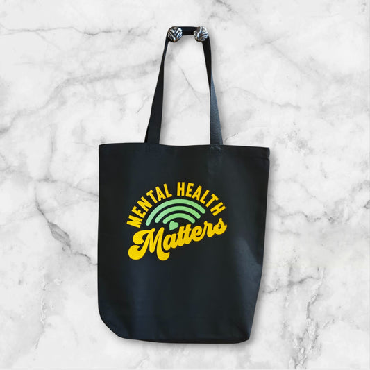 Black tote bag with 'Mental Health Matters' text and rainbow graphic on a marble background